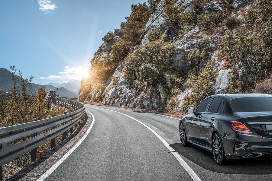 About Our Agency - Black Luxury Car Driving along a Mountain Highway, Rocky Ledge to the Right, Greenery to the Left